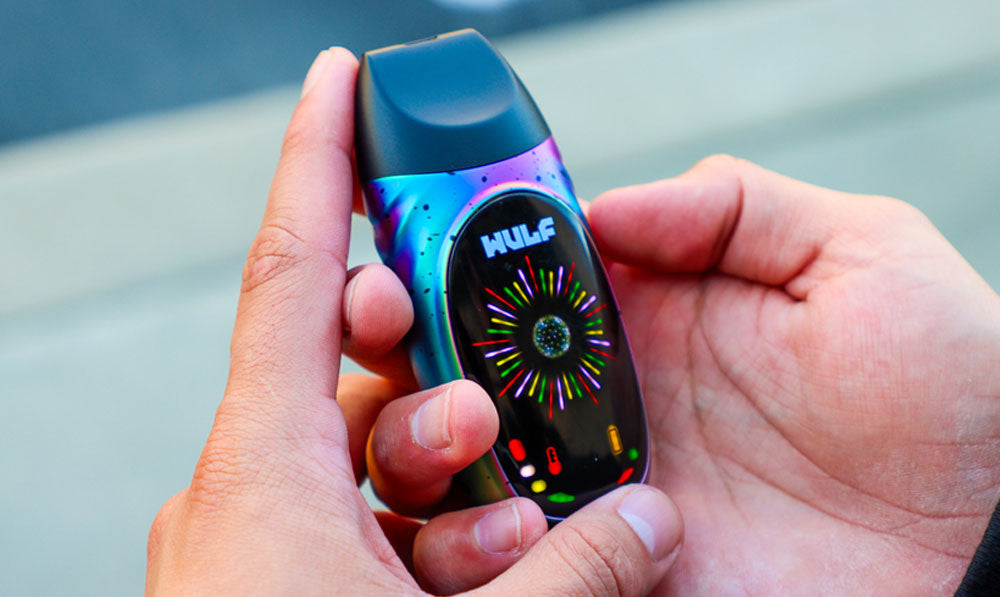 Hands holding a WULF FAZE vaporizer in iridescent blue and purple with a colorful starburst on its round display